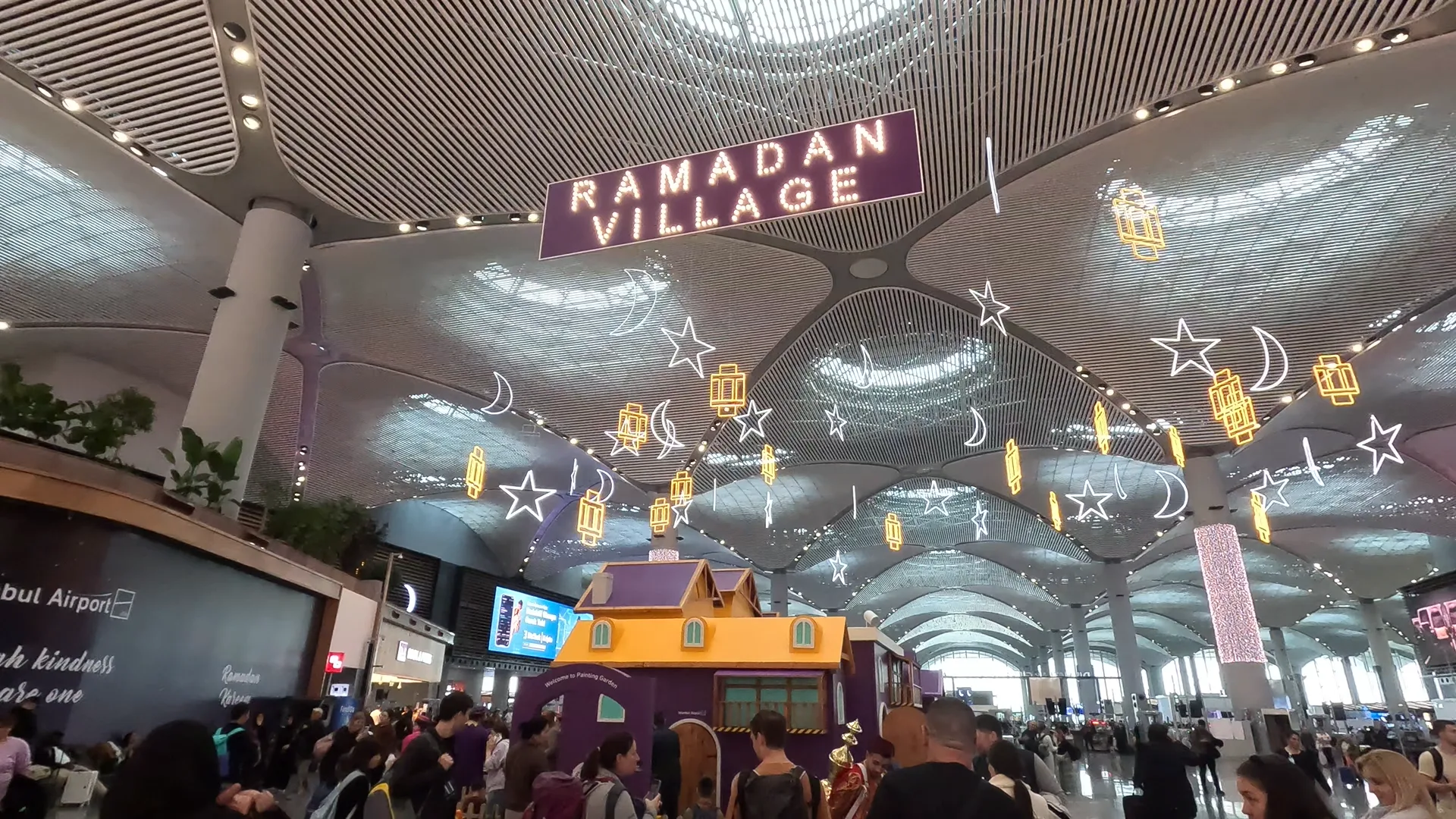 Busy crowd of passengers in front of the Ramadan Village at Istanbul Airport, various stalls and decorations of lit up stars and crescent moons.