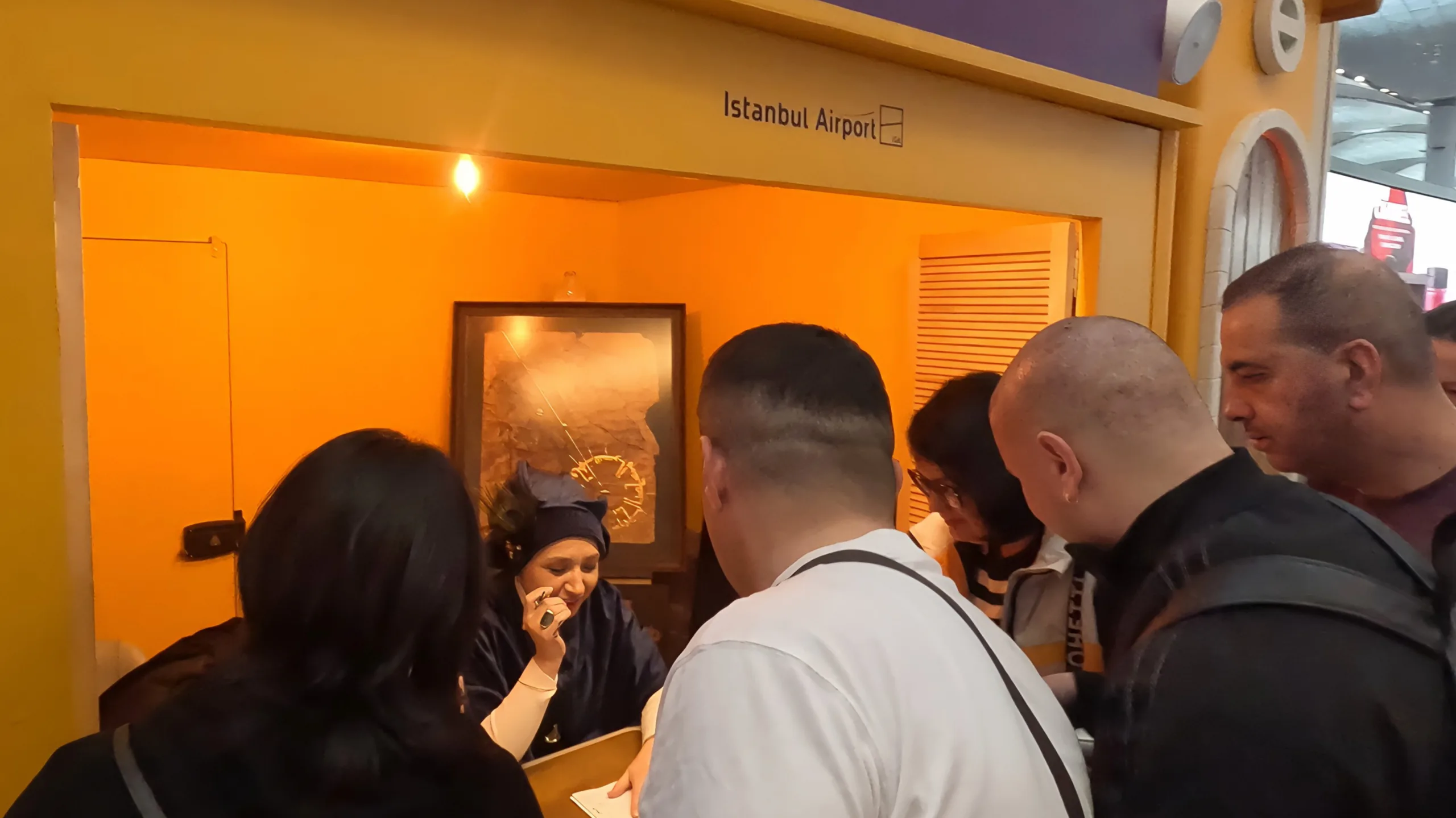 Crowd of people gathering around the Calligraphy Workshop stall and watching a female calligrapher write calligraphy at the Ramadan Village, Istanbul Airport.